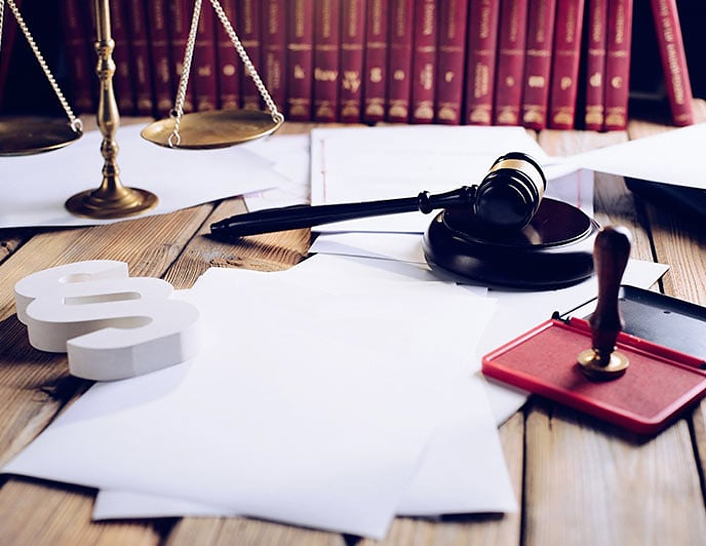Sur une table en bois, un marteau de juge, une balance dorée, un symbole paragraphe blanc, un tampon rouge et des documents devant des livres de droit rouges.