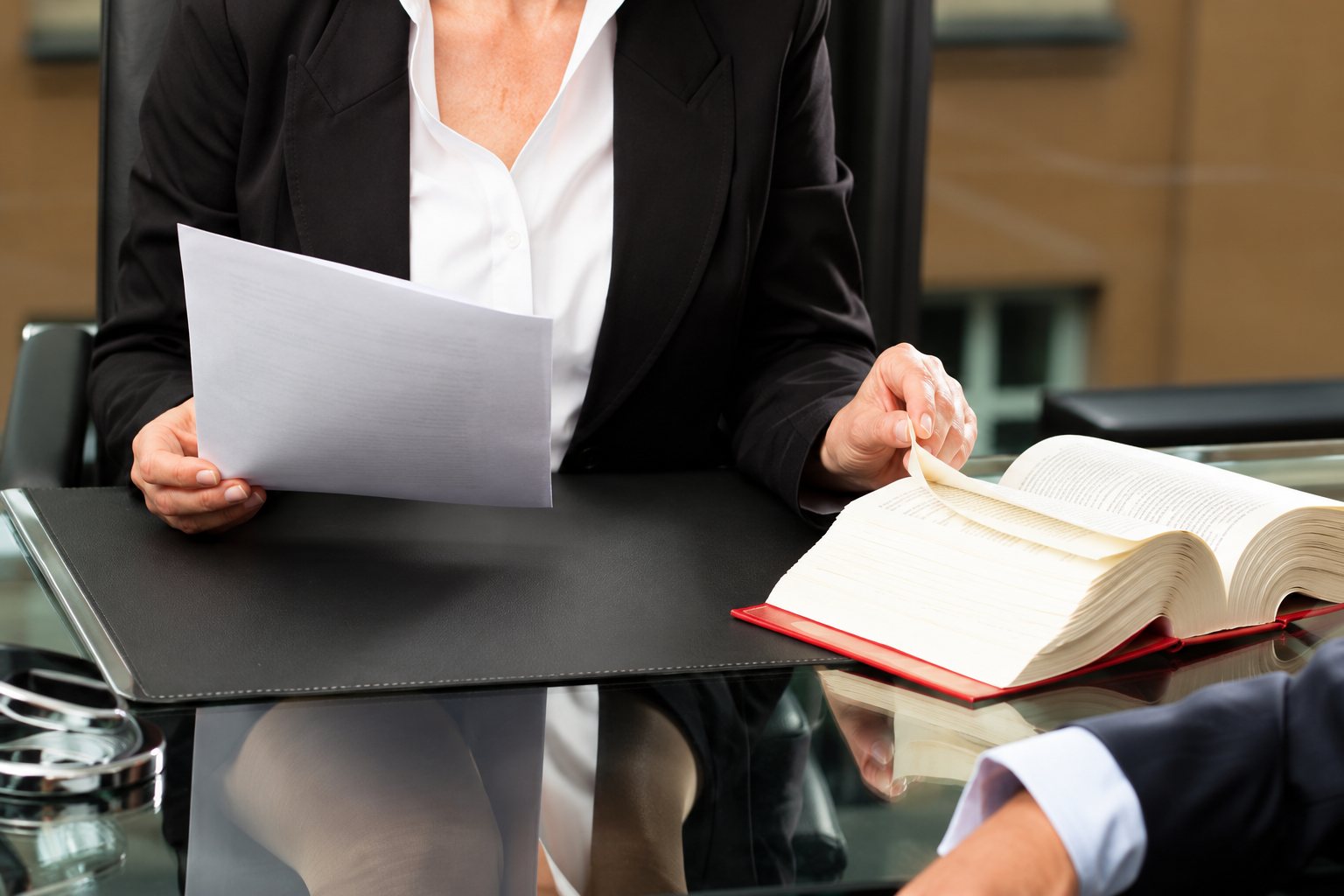 Une femme en tailleur noir tient une feuille de papier à une table, près d'un gros livre ouvert, dans un bureau.