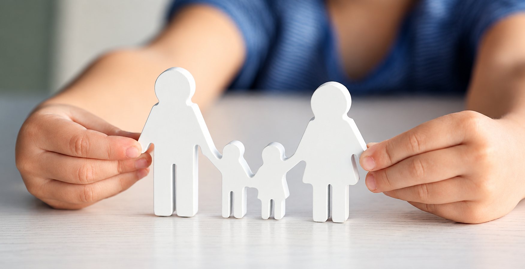 Deux mains d'enfant tiennent une famille de figurines blanches en bois, composée de deux parents et de deux enfants, posée sur une table claire.