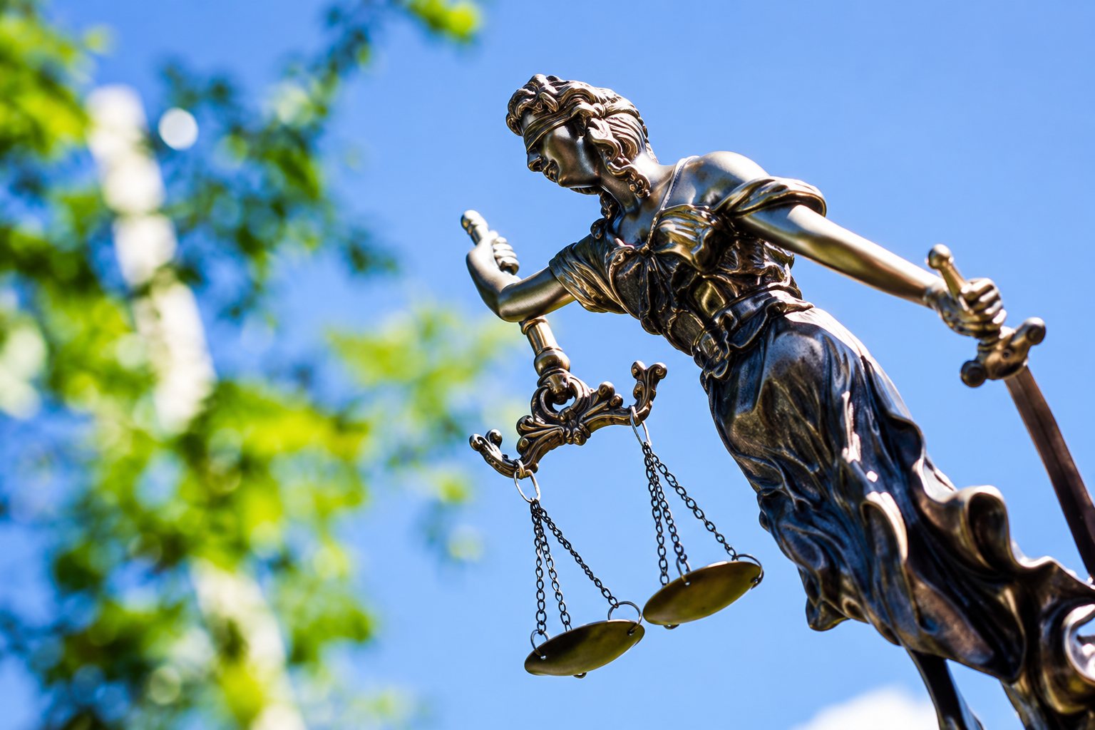 Statue en bronze de la déesse de la justice tenant une balance, photographiée en contre-plongée devant un ciel bleu et un feuillage flou.