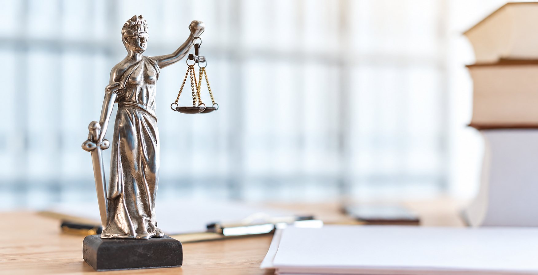 Une statuette argentée de la déesse de la justice, les yeux bandés, tient une balance et une épée, posée sur un bureau devant des livres empilés au fond clair et flou.