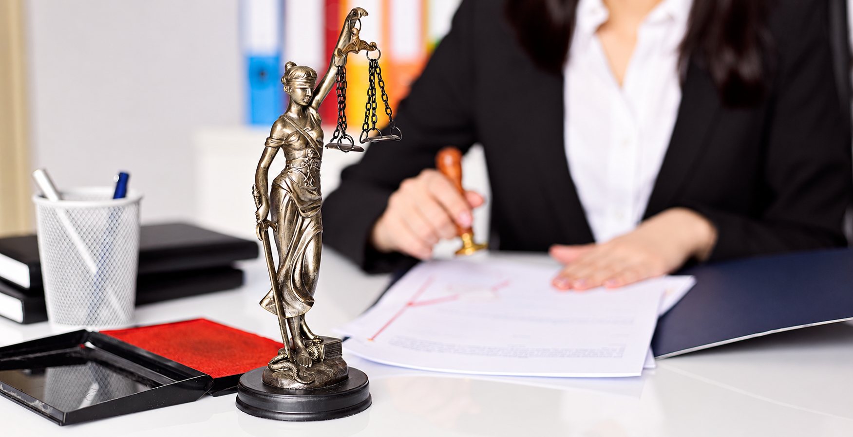 Une statuette de la justice aux yeux bandés tenant une balance est posée sur un bureau blanc, tandis qu'une femme en tailleur noir appose un tampon sur un document en arrière-plan flou.
